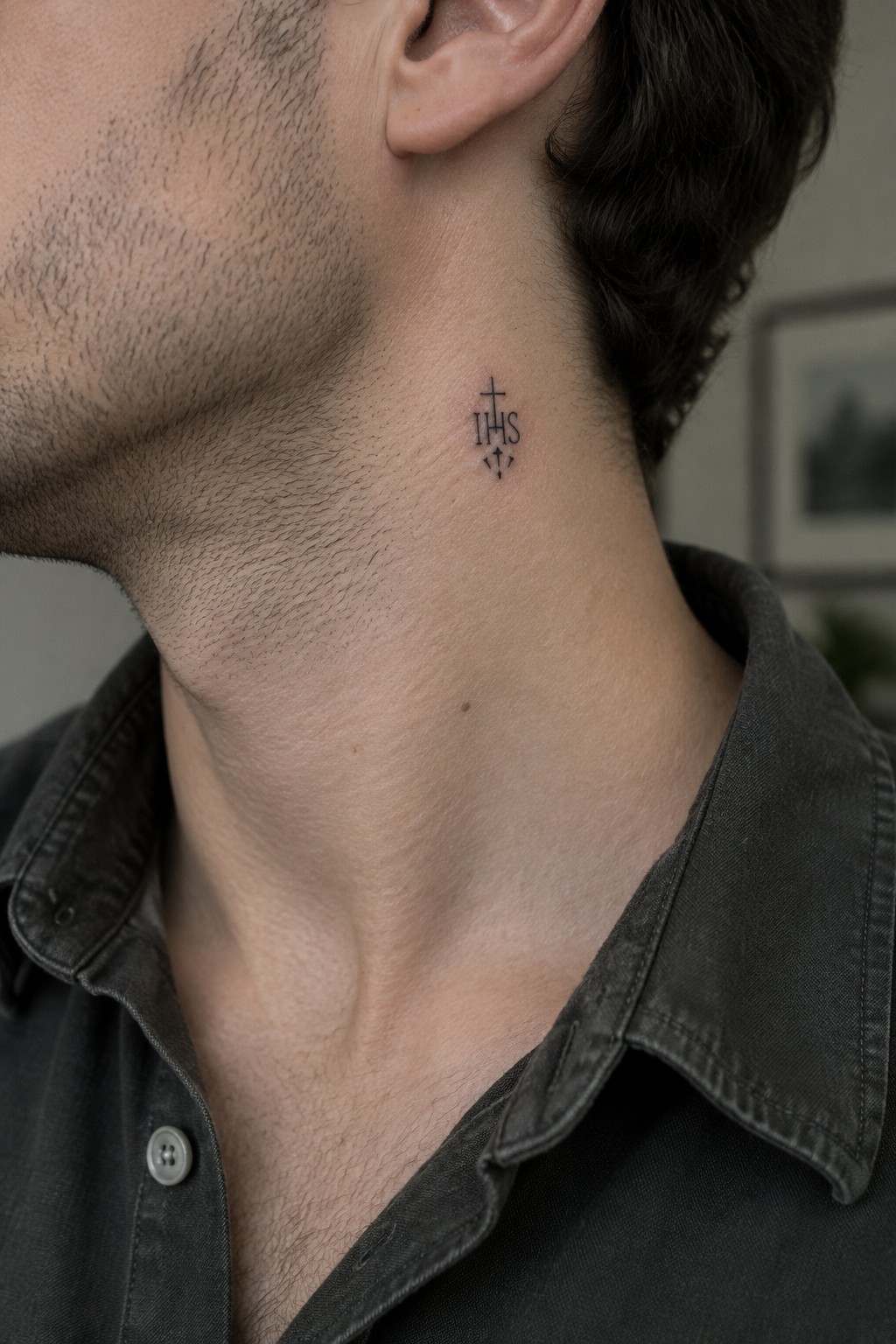Man's neck with small black IHS cross tattoo above dark button-up shirt collar