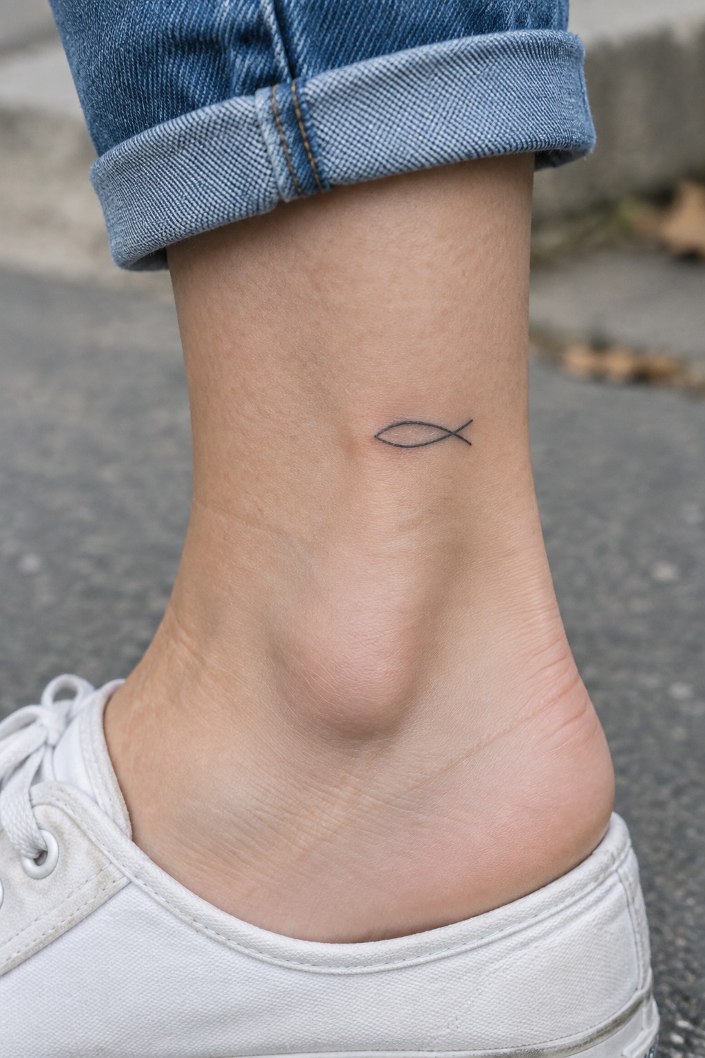 Small black fish tattoo on ankle above white sneaker and cuffed blue jeans