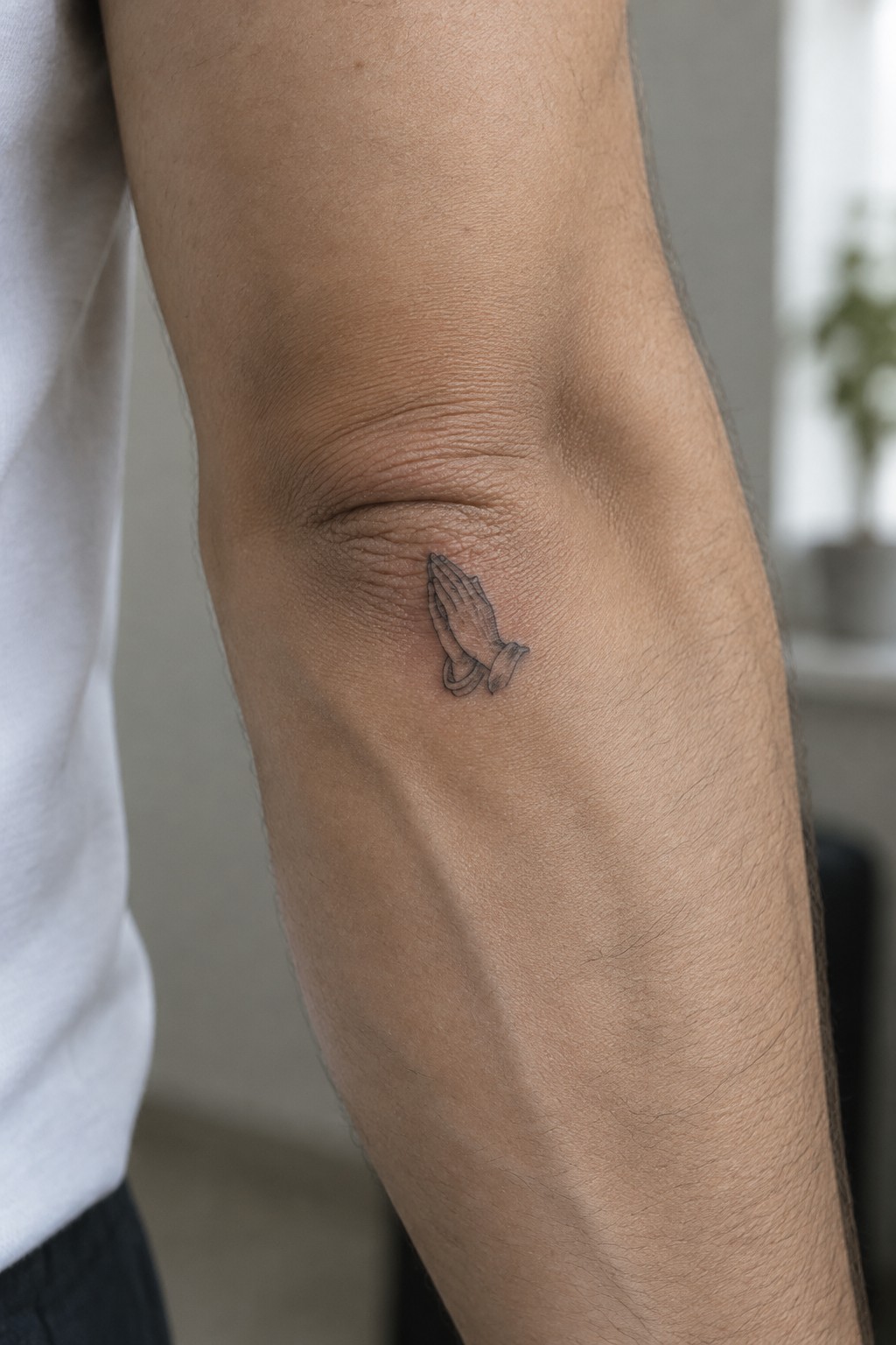Small black line art tattoo of praying hands on the inner elbow of a man's arm