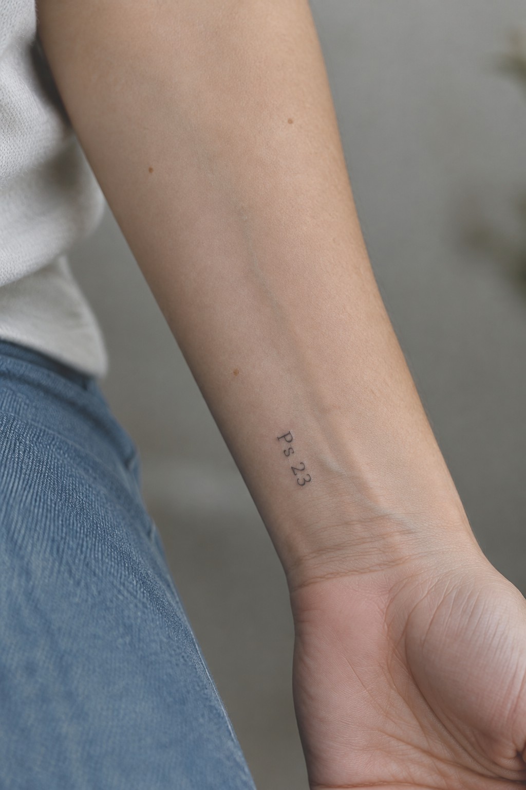 Small black text tattoo reading Ps 23 placed vertically on the inner forearm of a person in a white shirt and blue jeans.