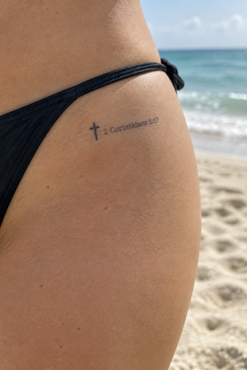 Small black cross tattoo with 2 Corinthians 5:17 text on the hip area of a person at the beach