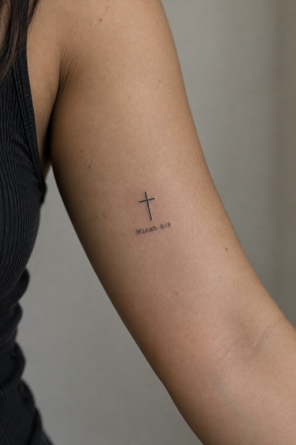 Small black cross and Micah 6:8 tattoo on upper arm near black tank top
