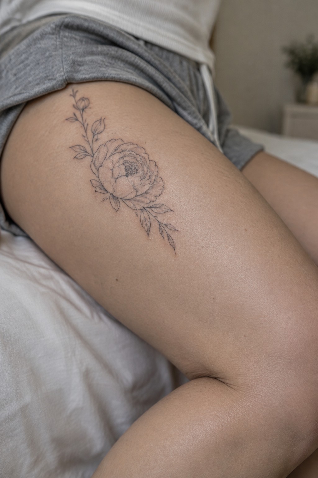 Line art peony tattoo with leaves on thigh beside grey shorts on white fabric