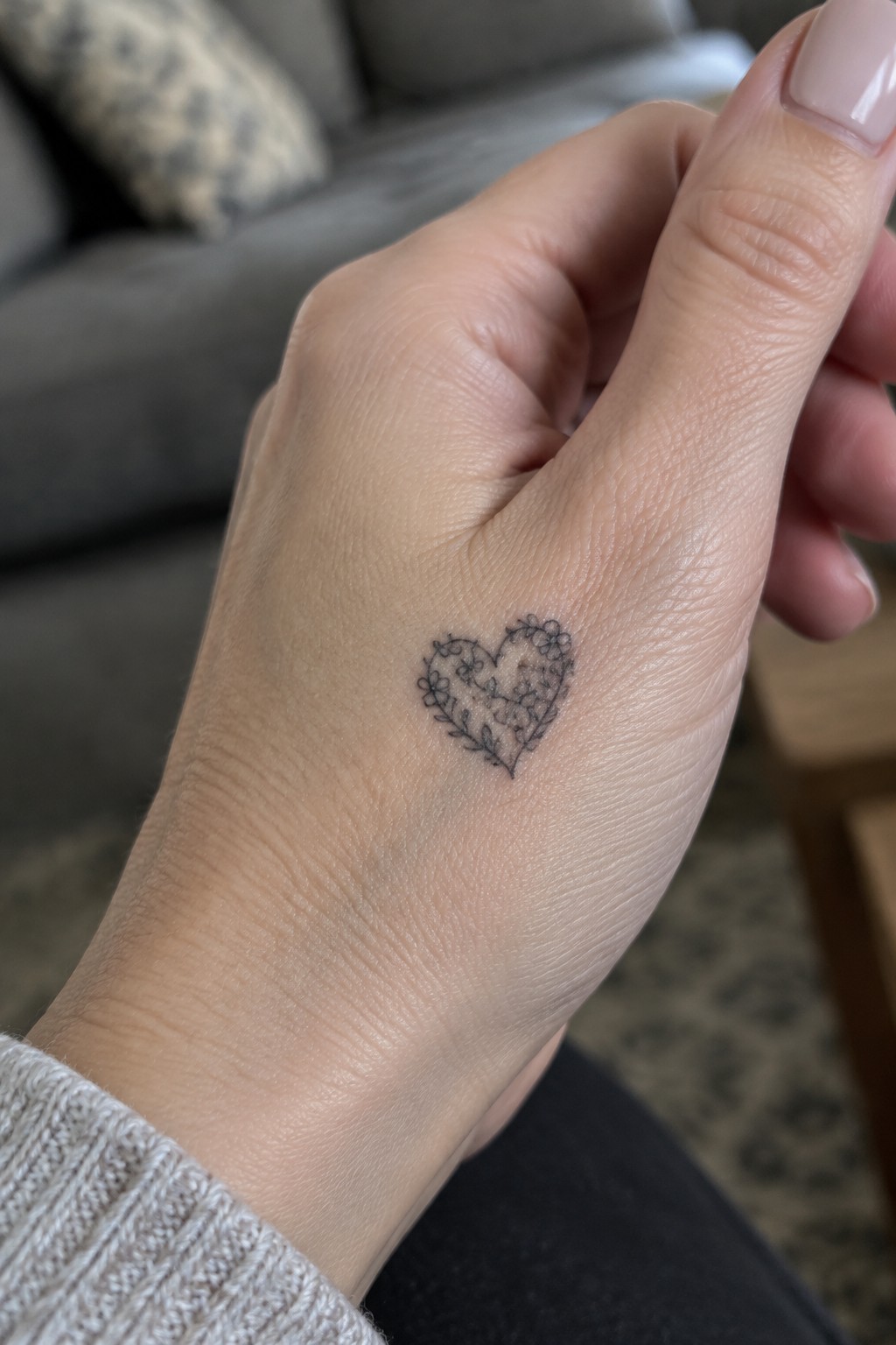 Hand with floral heart tattoo, light knit sweater, and pink manicured nails