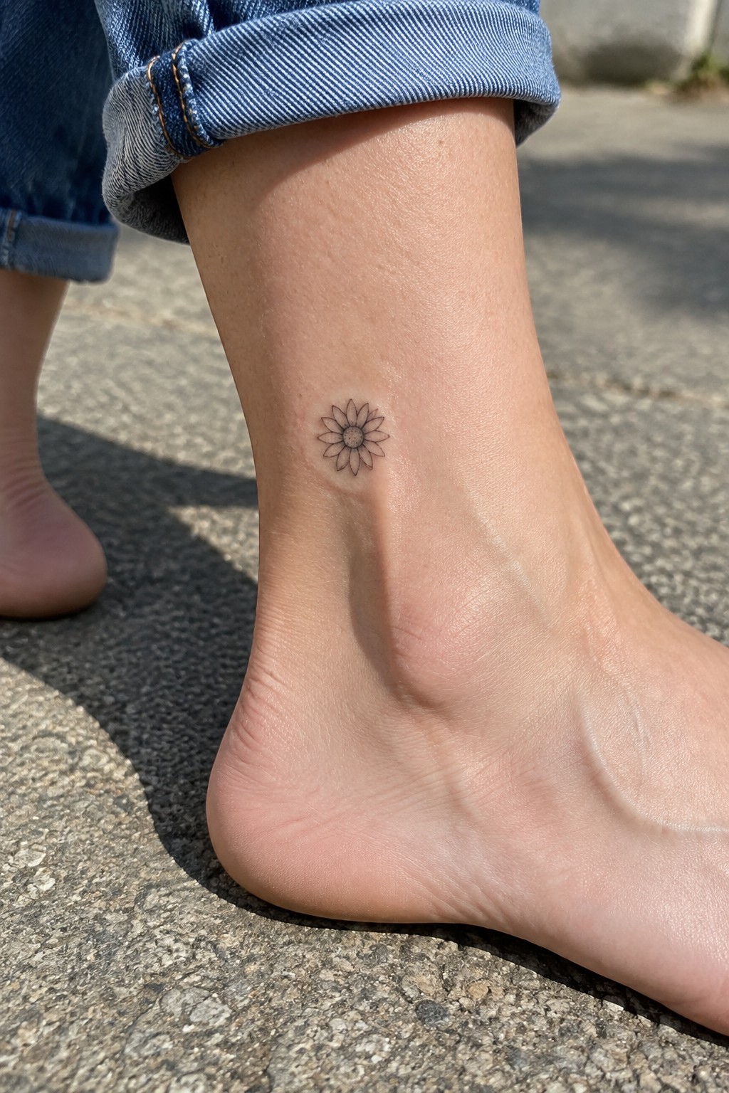 Small daisy tattoo on ankle below cuffed blue jeans on pavement