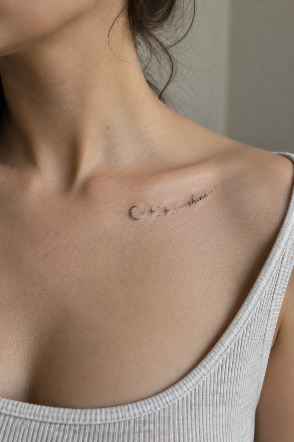 Fine-line tattoo of a crescent moon, two stars, and the word “shine” along a woman’s collarbone.