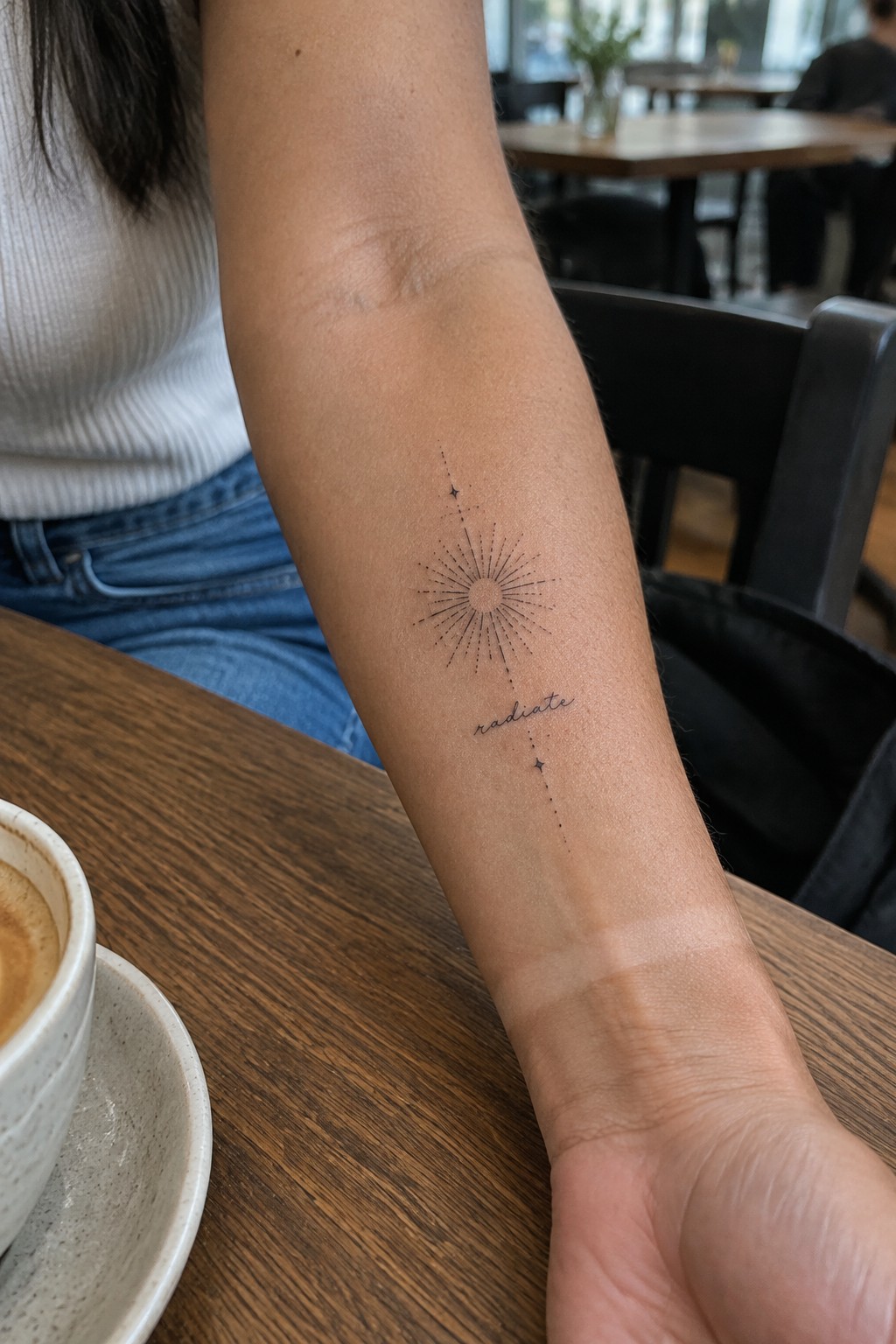 Fine-line sunburst tattoo with tiny stars and the word “radiate” on the inner forearm.