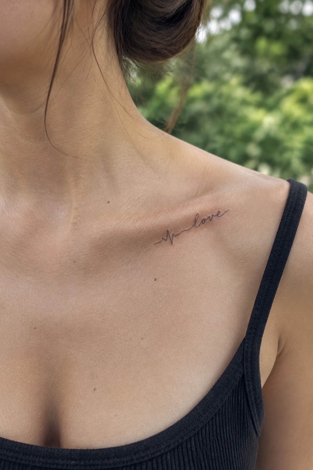 Fine-line heartbeat tattoo leading into the word “love” on a woman’s collarbone.