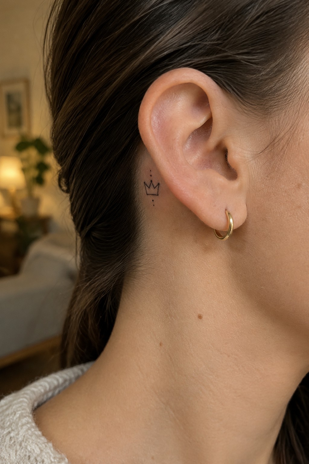 Small fine-line crown tattoo behind a woman’s ear, with tiny dots above and below it.