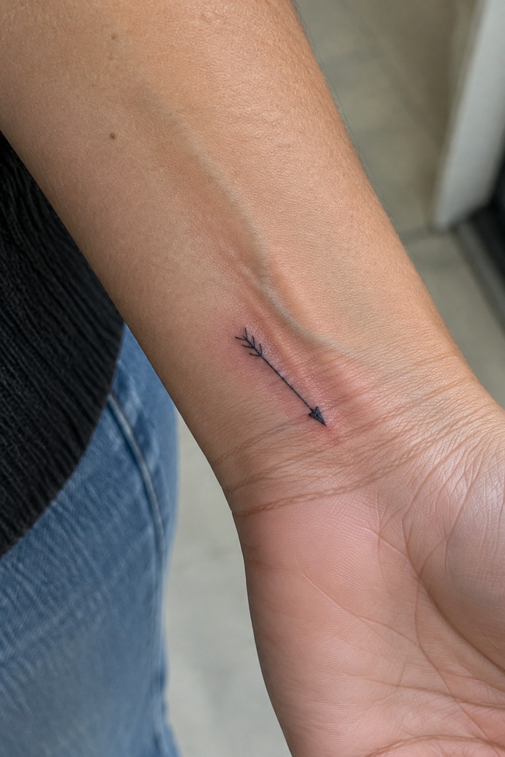 Fine-line black arrow tattoo with small feather details on the inner wrist.