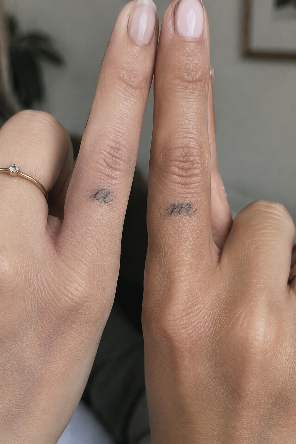 Two hands with small fine-line script tattoos on the fingers, one showing a lowercase “a” and the other a lowercase “m.”