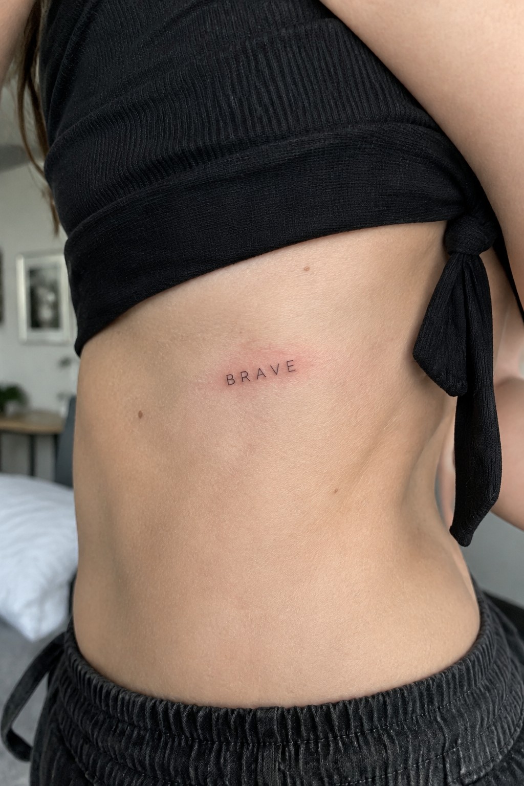 Small black text tattoo spelling the word brave on the side torso above dark pants