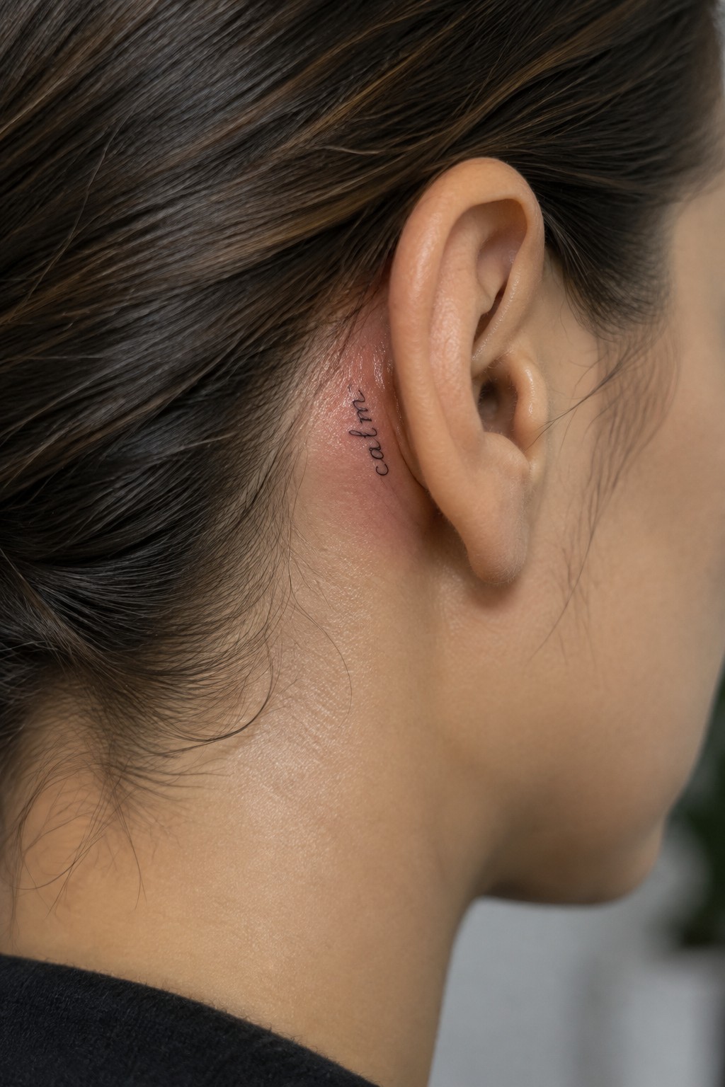 Small 'calm' tattoo in script behind ear on reddened skin with brown hair