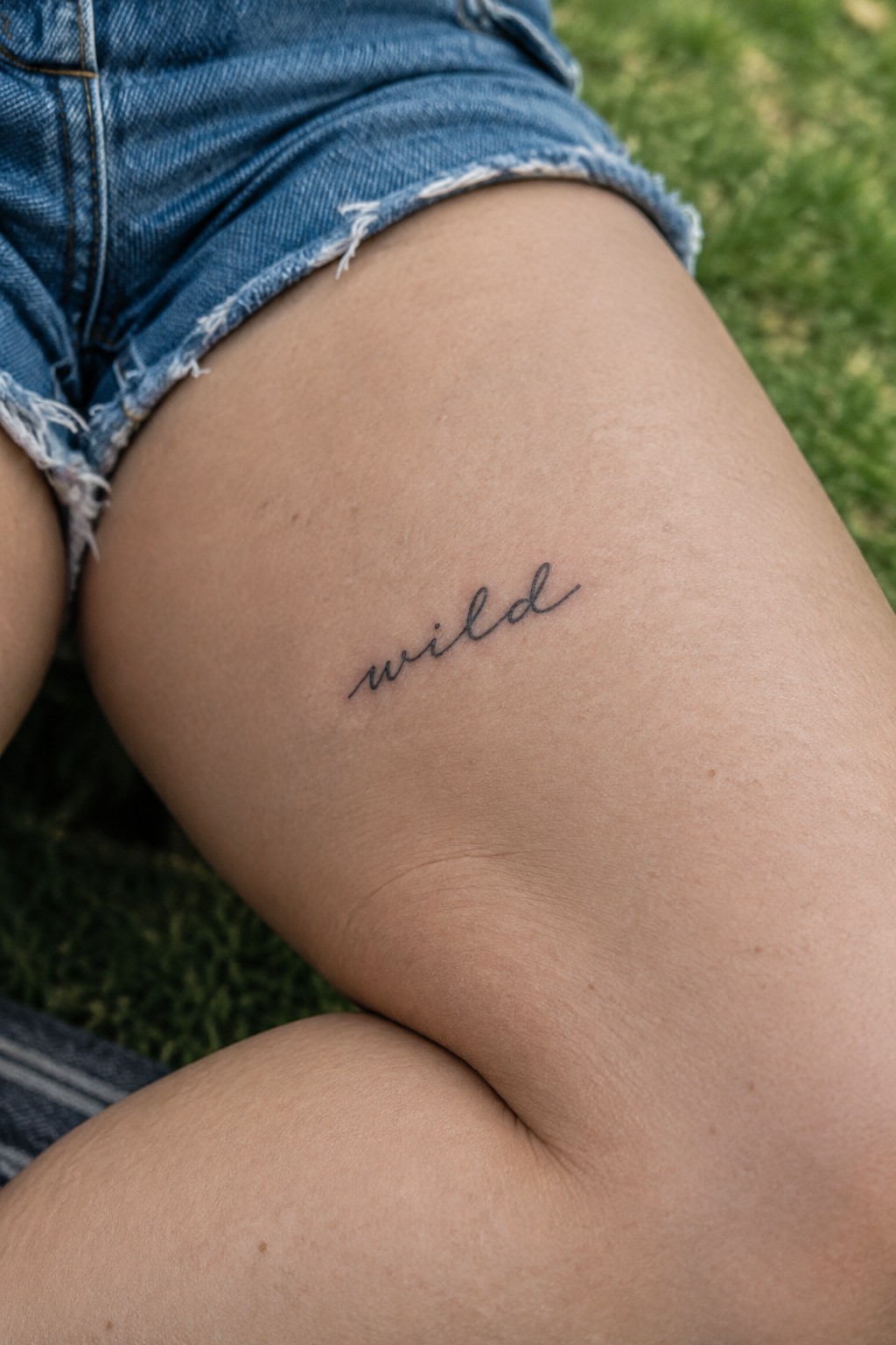 Small cursive tattoo of the word wild on the upper thigh under denim shorts.