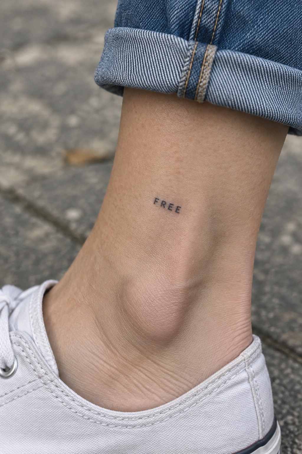 Close-up of ankle with black 'FREE' tattoo, rolled blue jeans, white sneaker.