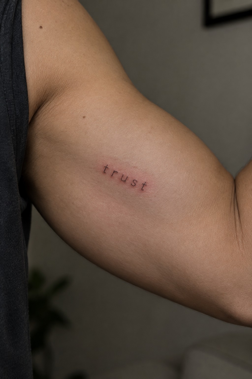 Small lowercase trust tattoo in thin black lettering on the inner upper arm with light redness around the fresh ink