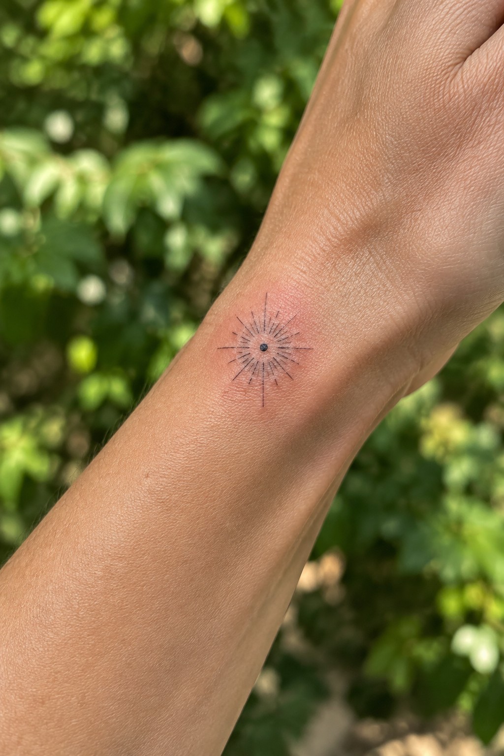 Black starburst tattoo on light-skinned inner wrist with blurred green foliage behind.