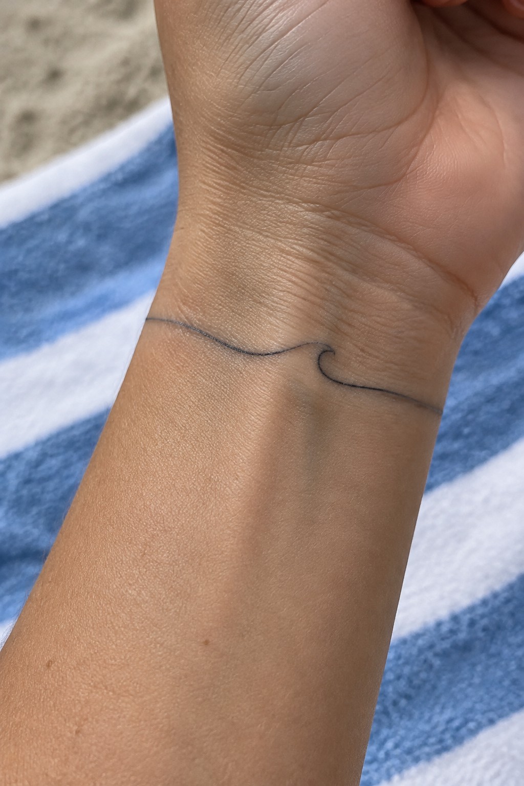 Minimalist black wave tattoo on wrist over blue striped beach towel