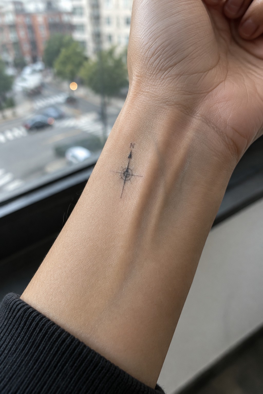 Black compass tattoo on wrist above black sweater cuff, blurred city view behind.