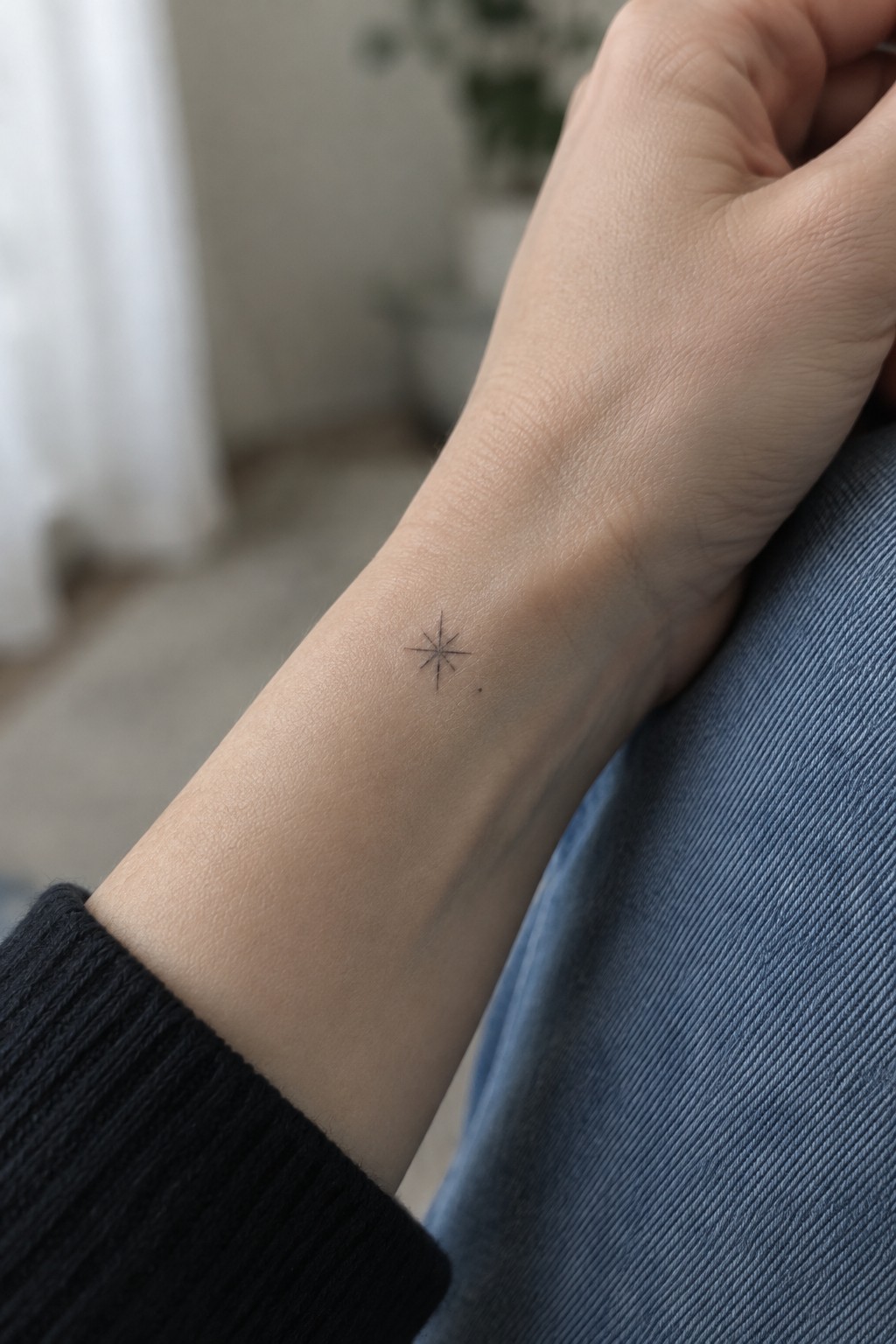 Small black fine line star tattoo centered on the inner wrist of a light skinned arm resting on blue fabric.