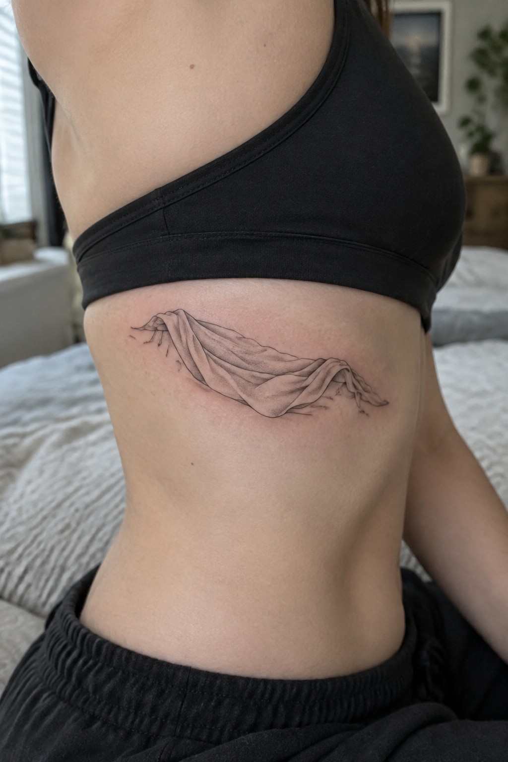 Fine line black and grey tattoo of a draped cloth across the side torso representing a resurrection symbol