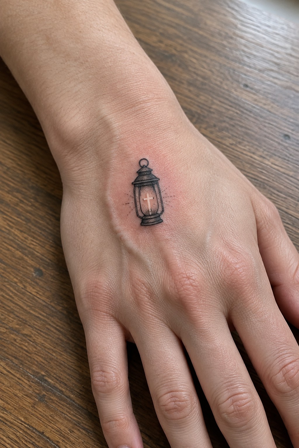 Minimalist black ink tattoo of a lantern with a cross inside on the back of a hand