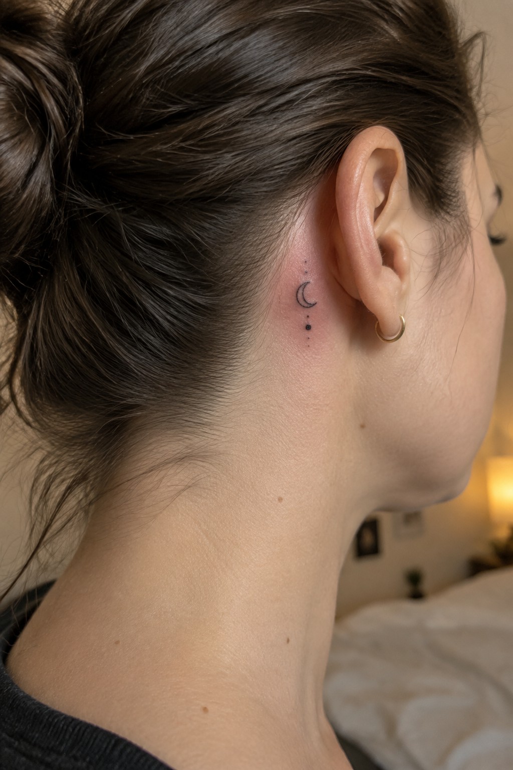 Small black crescent moon tattoo with a line of dots behind a woman’s ear.