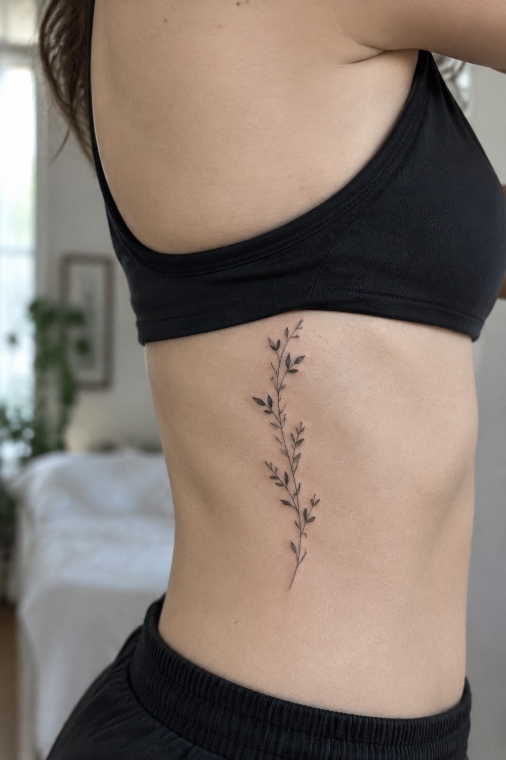 Fine-line leafy branch tattoo running vertically along a woman’s side below a black sports bra.