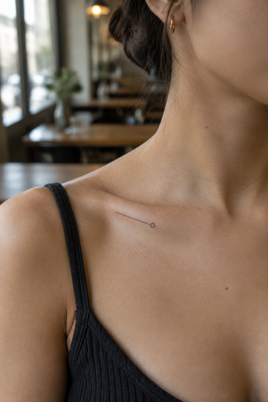 Fine line tattoo of a thin diagonal line ending in a small open circle on a woman’s collarbone.