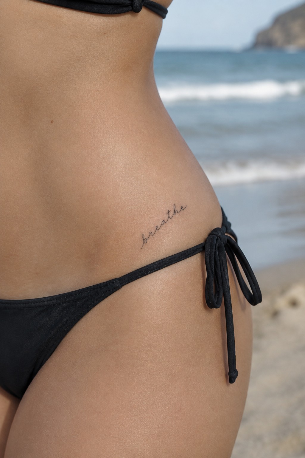 Small fine-line “breathe” script tattoo on the lower abdomen above the bikini line.