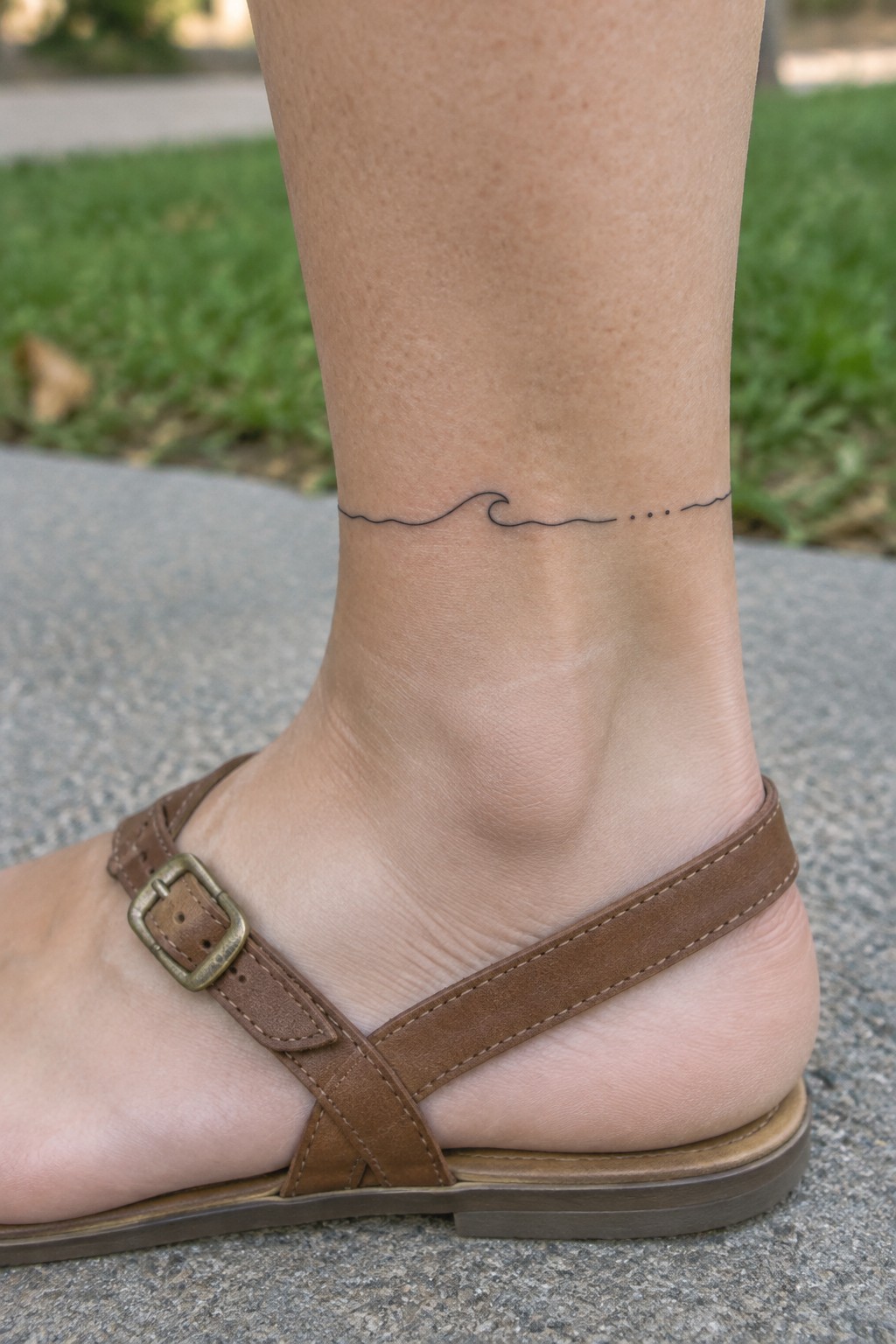 Fine-line wave tattoo with three small dots wrapping around a woman’s ankle above a brown sandal.