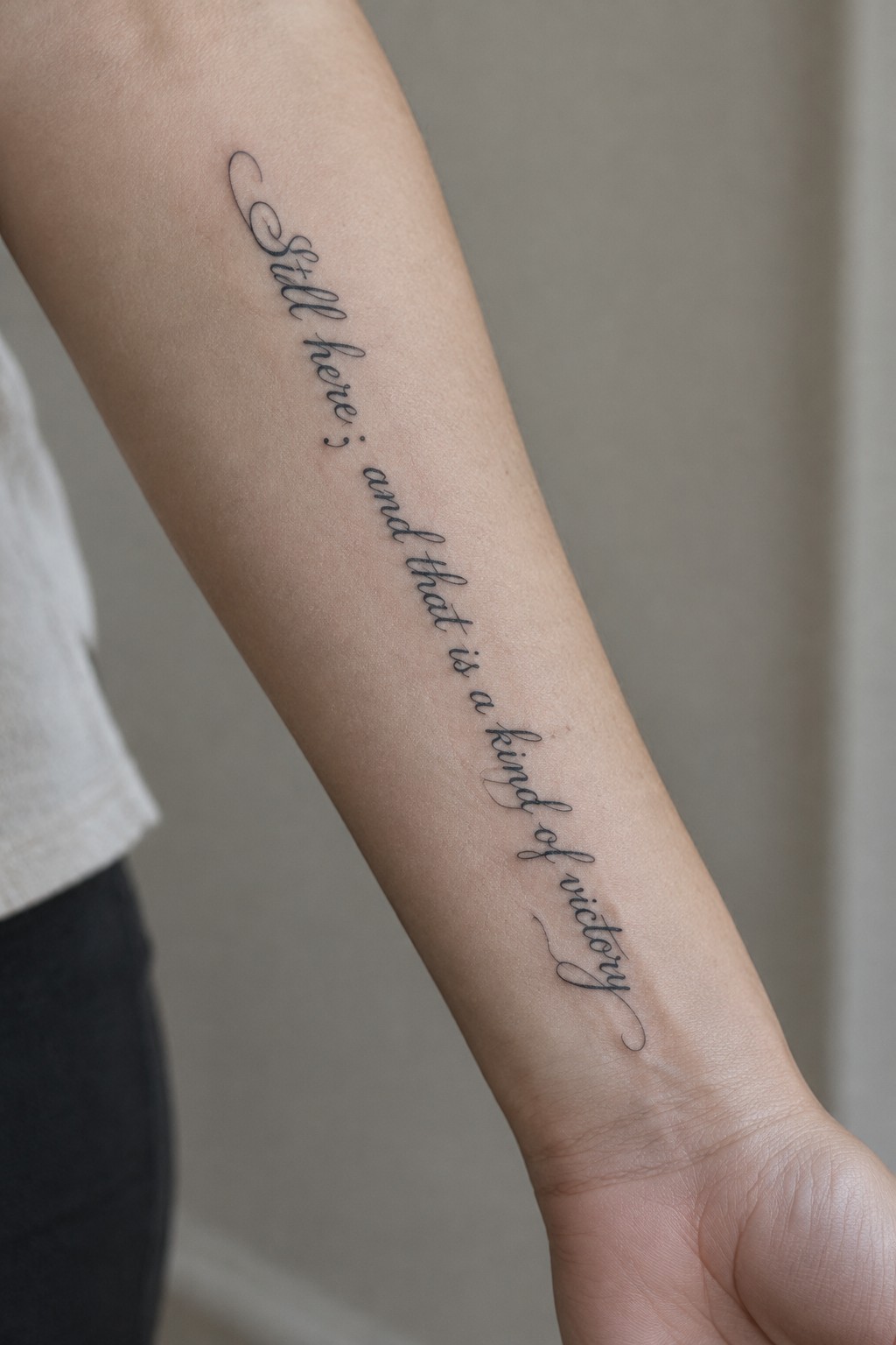 Fine black script tattoo reading “Still here; and that is a kind of victory” along the inner forearm.