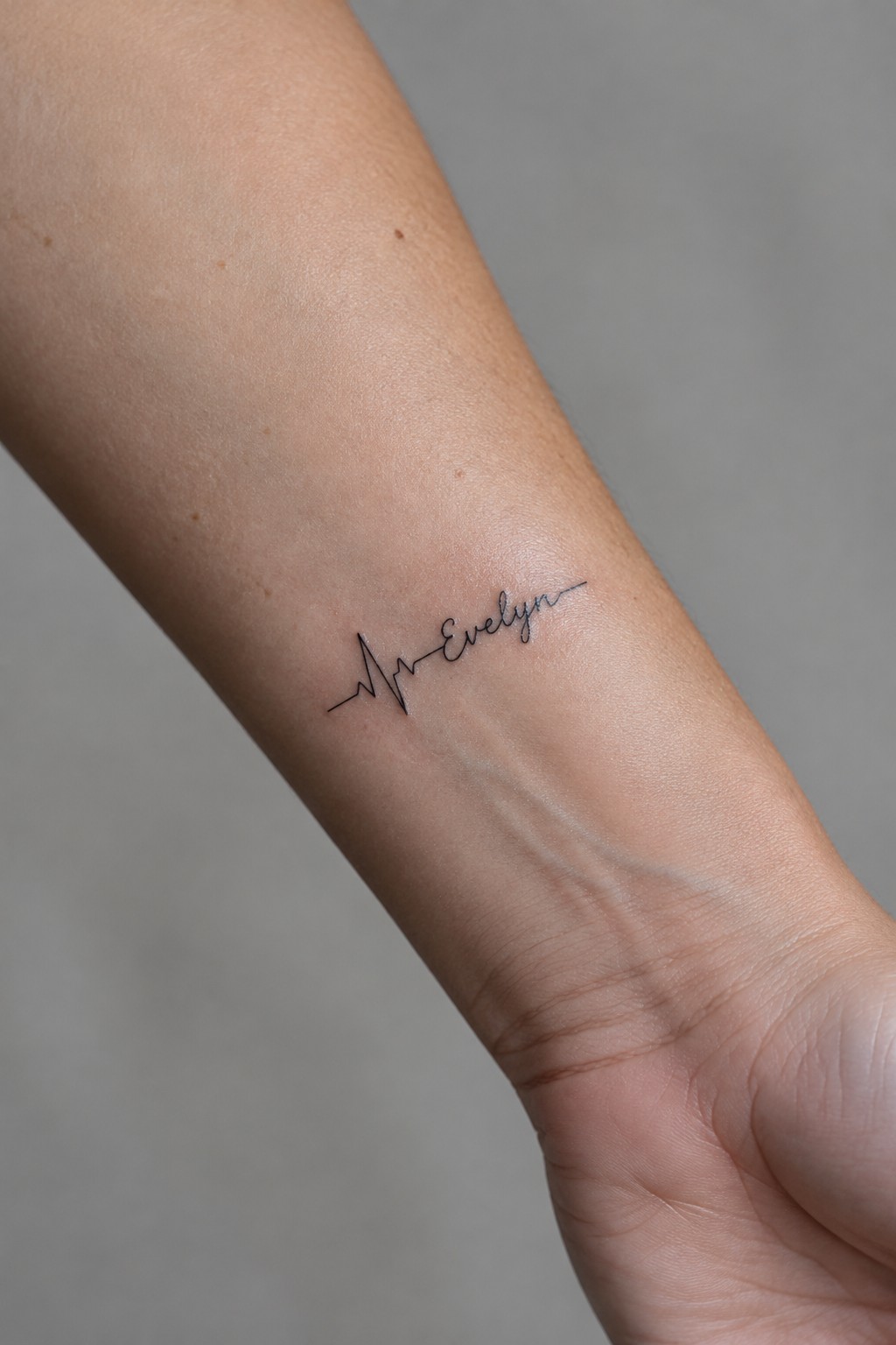 Fine-line black tattoo of the name Evelyn connected to a heartbeat line on the inner forearm.