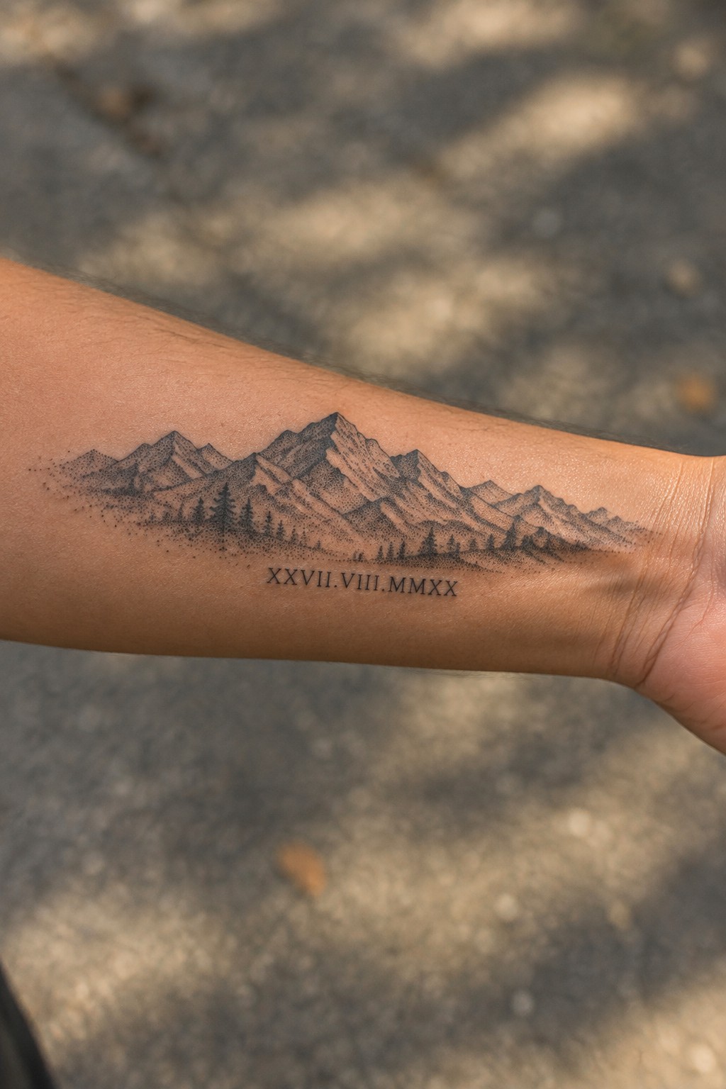 Fine-line mountain range tattoo with small trees and a Roman numeral date across the inner forearm.