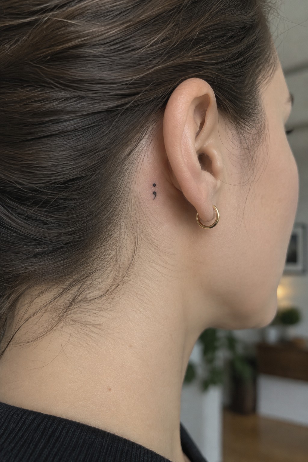Small black semicolon tattoo behind a woman’s ear, beside a gold hoop earring.
