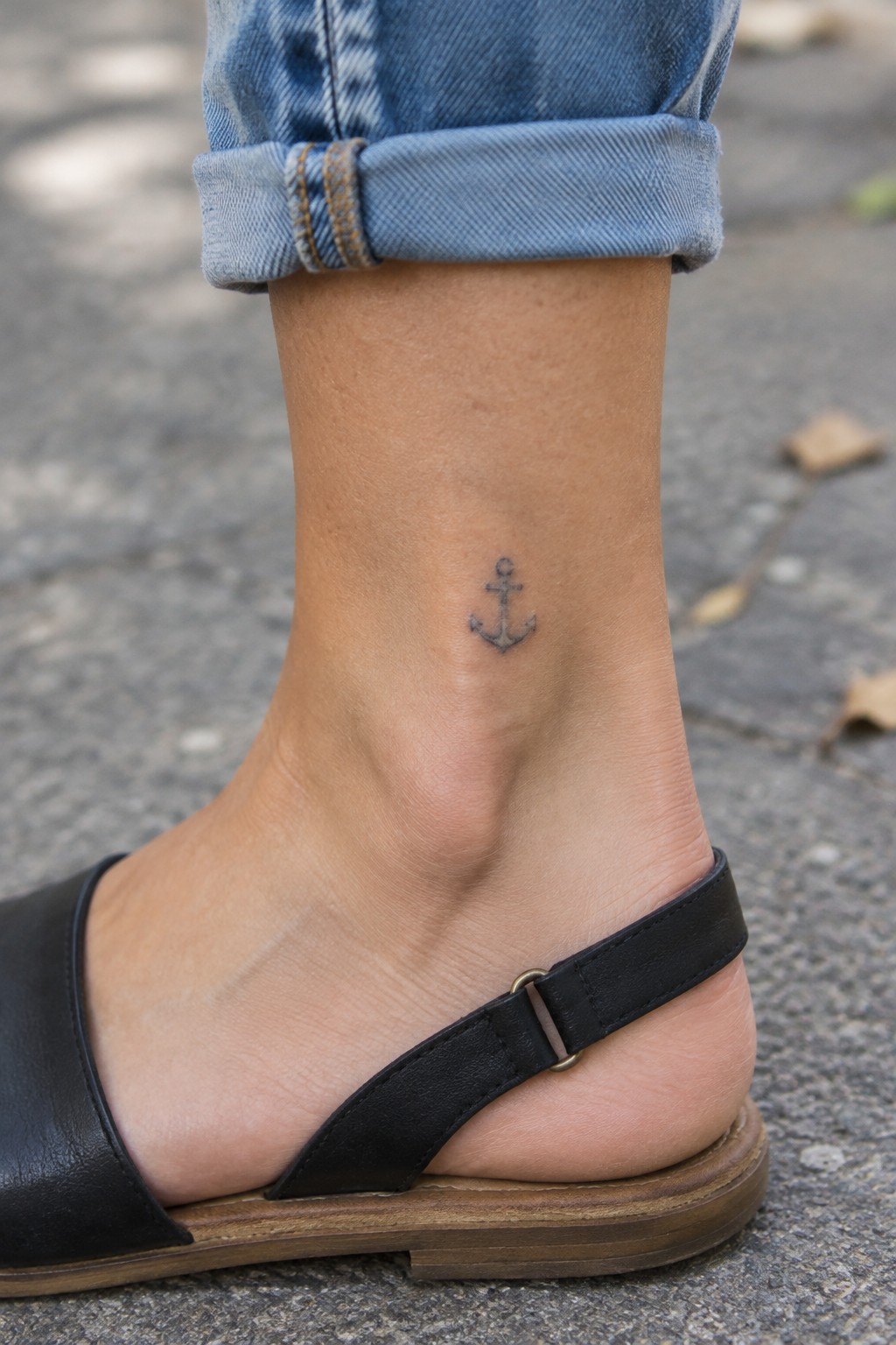 Small black anchor tattoo on the outer ankle above a black sandal, with rolled-up jeans.