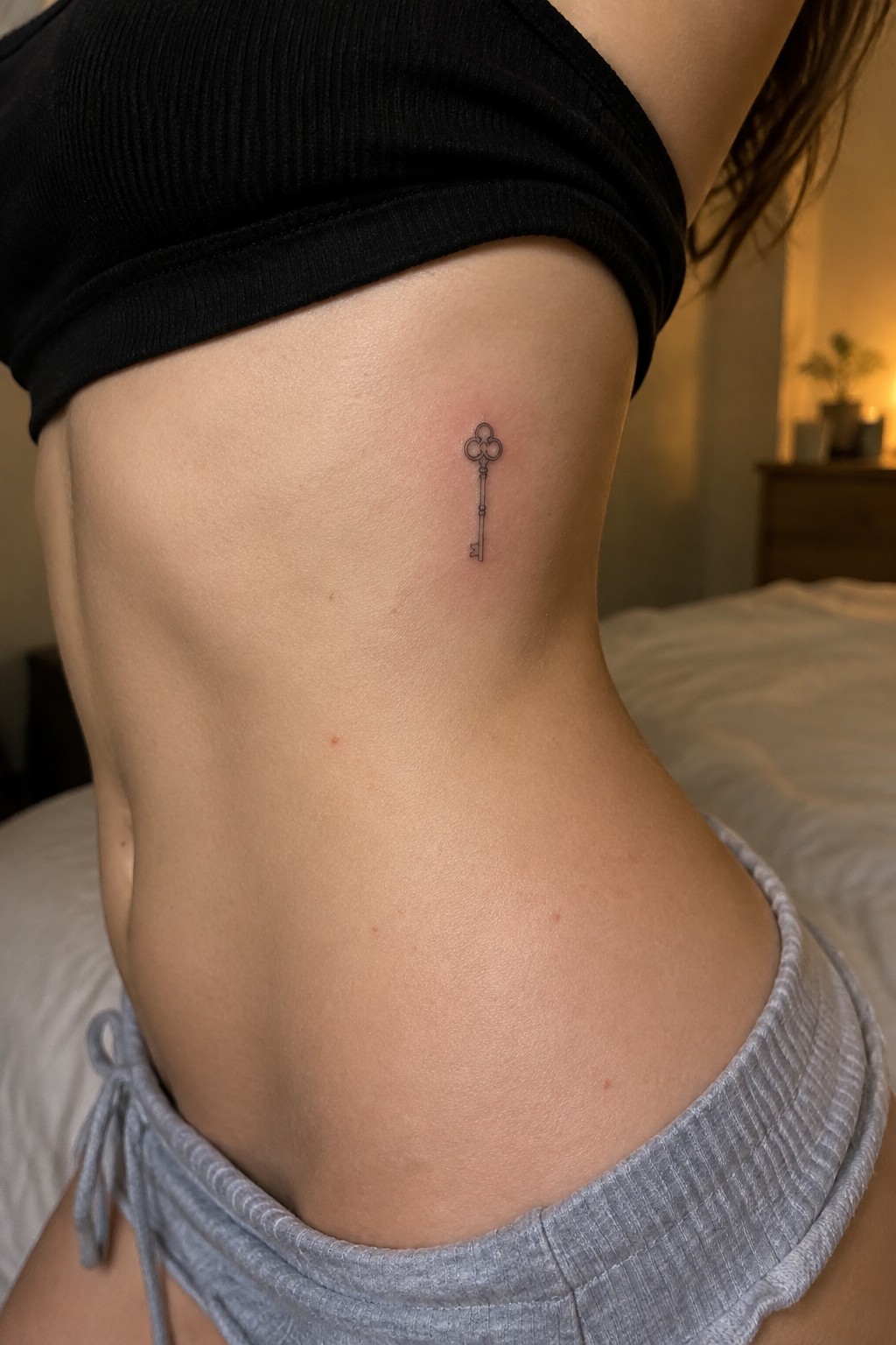 Small fine-line key tattoo on the side of the ribcage beneath a black top.