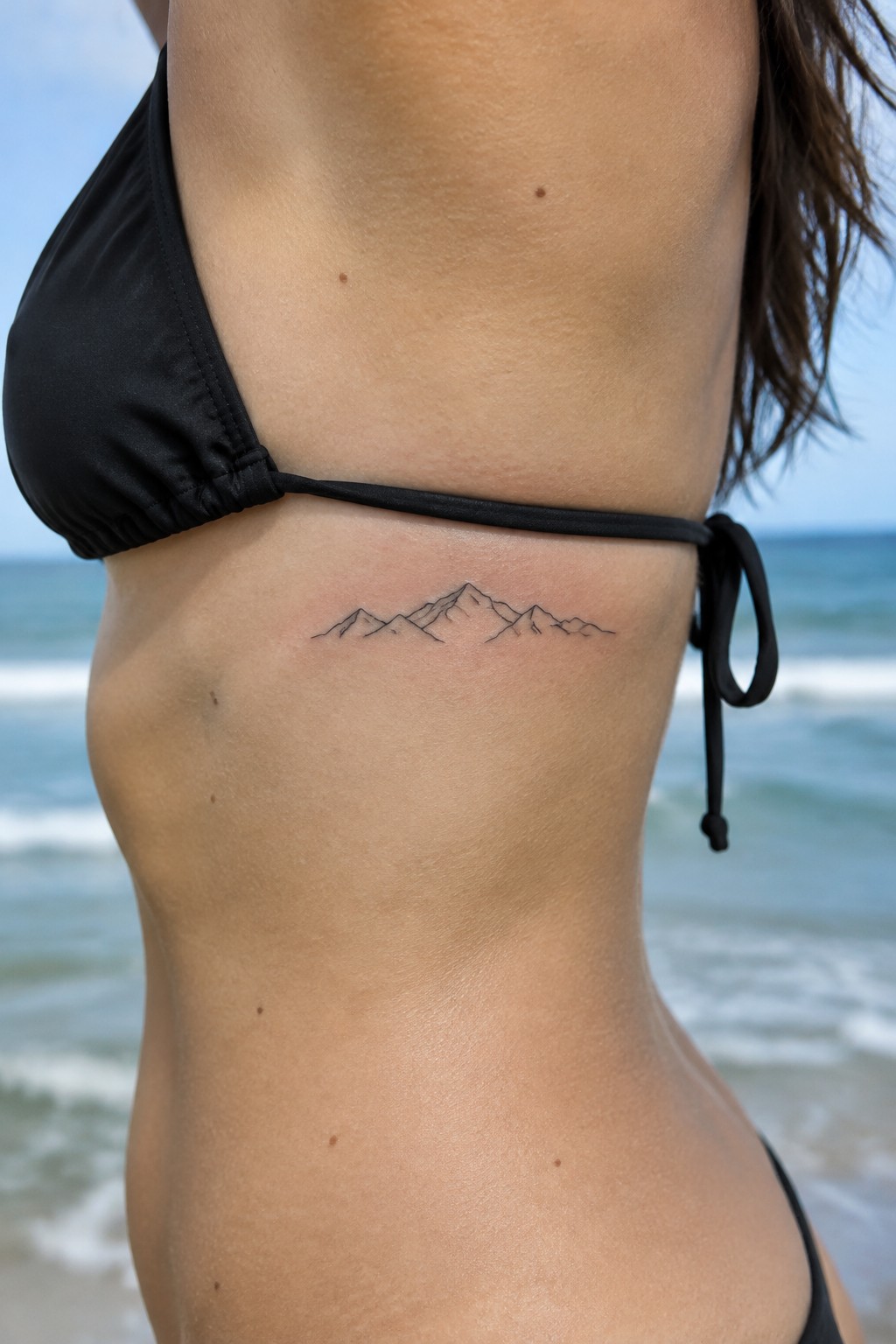Fine-line mountain range tattoo along a woman’s ribs beneath a black bikini top at the beach.