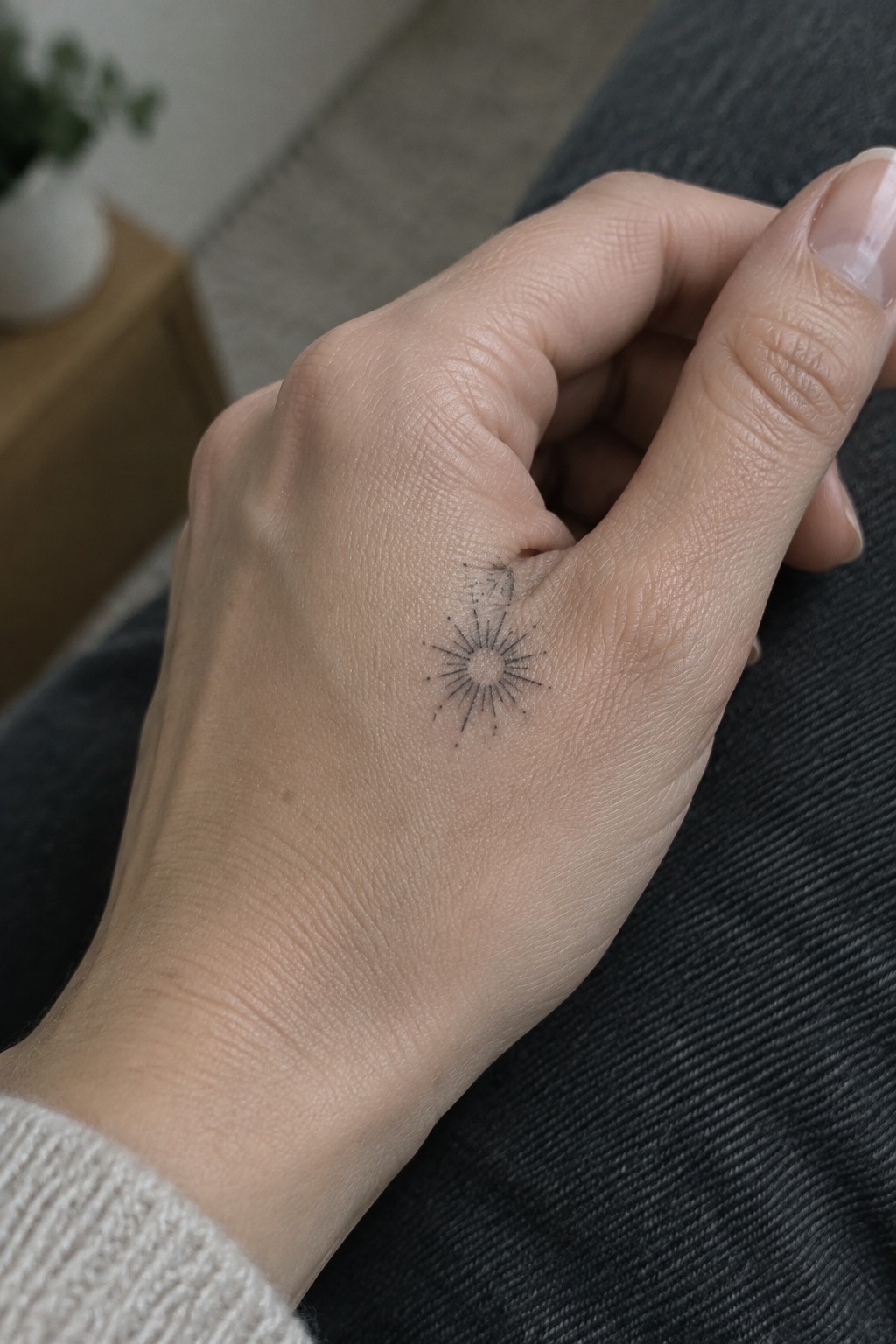 Small fine-line sunburst tattoo with scattered dots on the back of a hand near the thumb.
