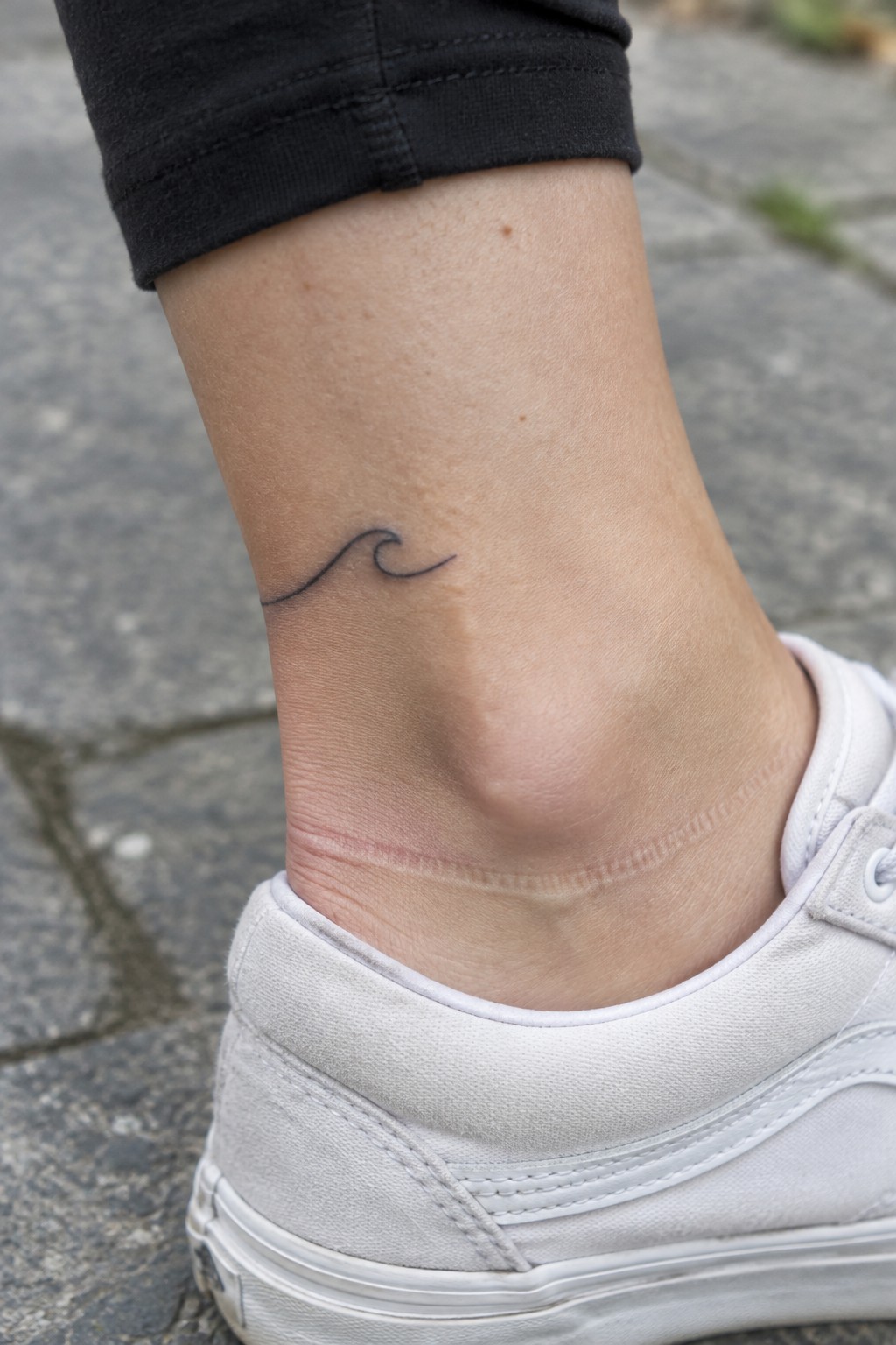 Small black fine-line wave tattoo on the side of an ankle above a white sneaker.