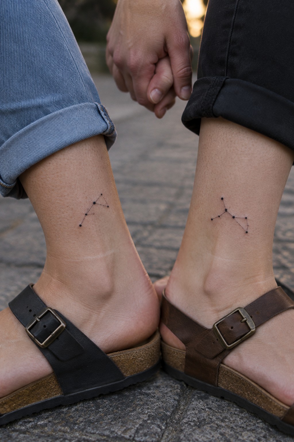 Two people hold hands with small black constellation tattoos above their ankles, paired with rolled-up jeans and sandals.