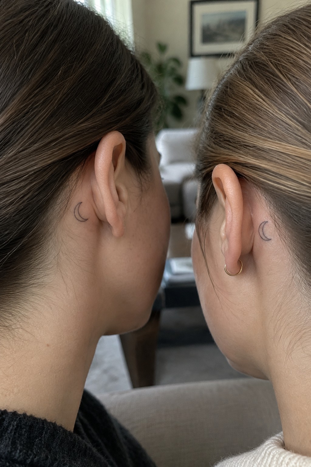 Two women showing matching small crescent moon outline tattoos behind their ears, one with a gold hoop earring.