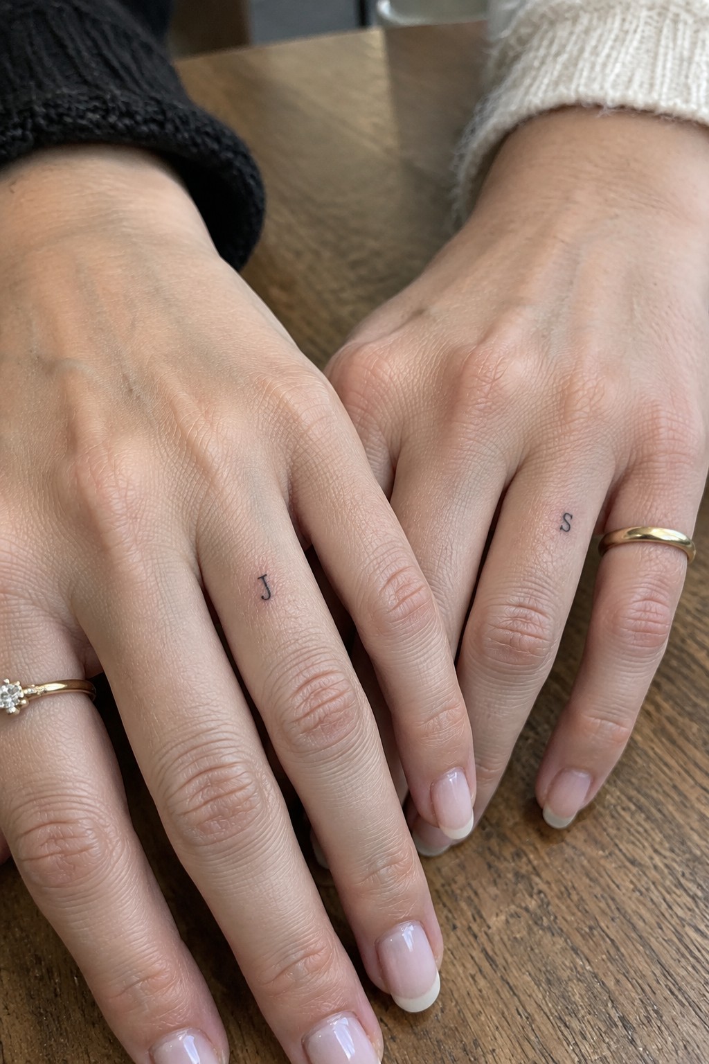 Two hands with small black initial tattoos on the fingers and gold rings.