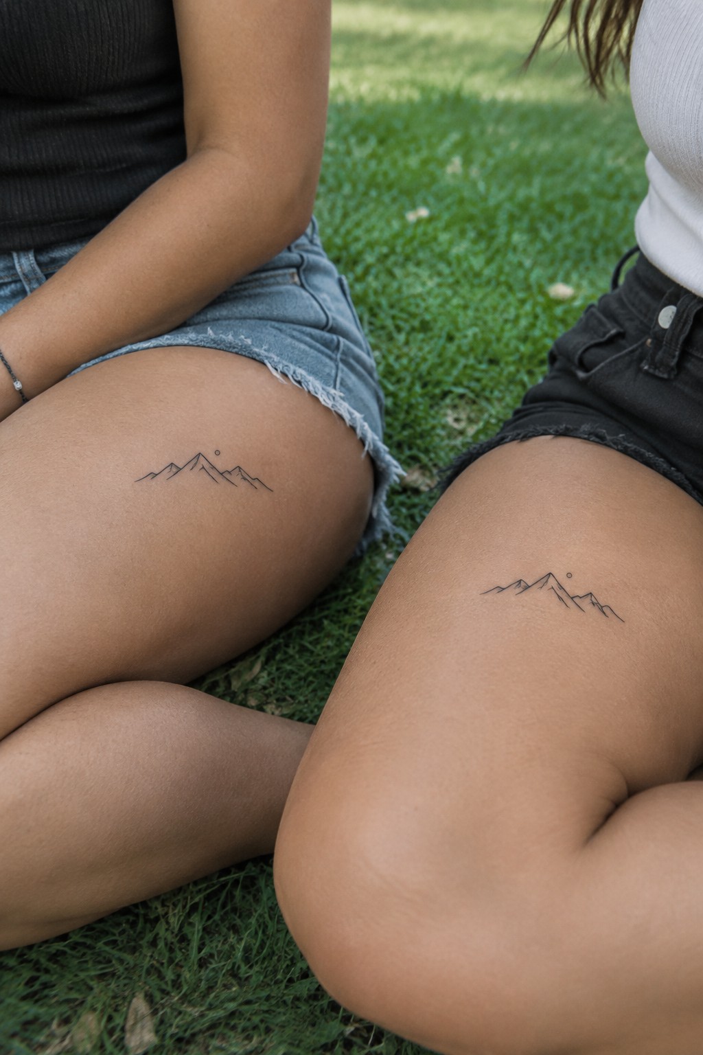 Two women sitting on grass with matching small fine-line mountain tattoos on their upper thighs.