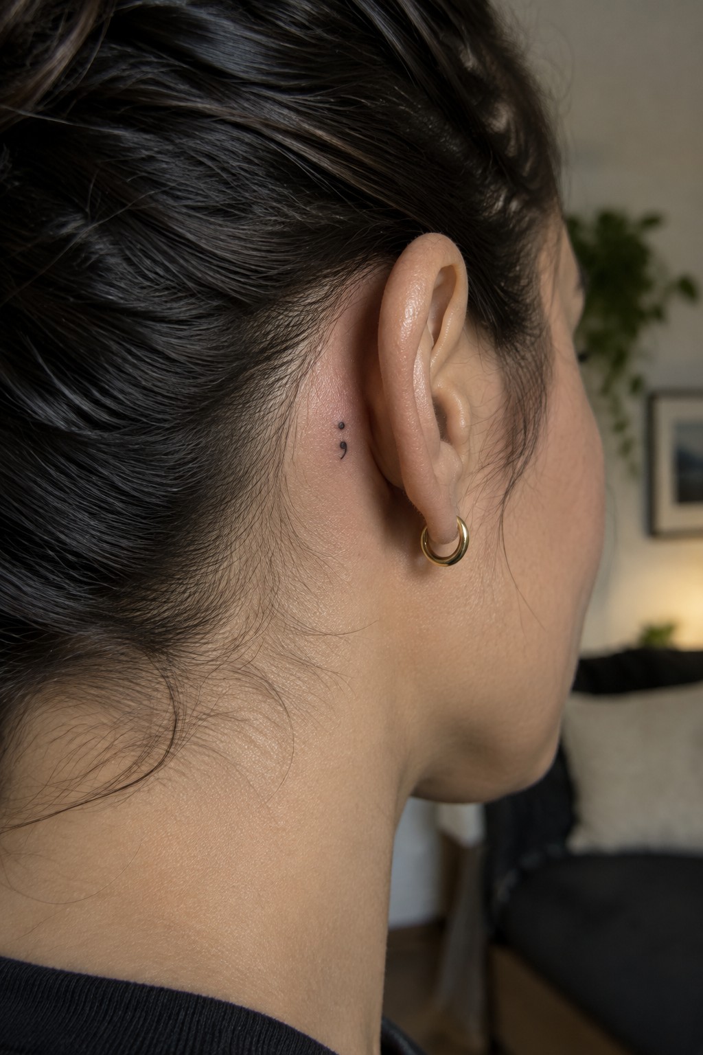 Close-up of a small black dot and curved tattoo behind a woman’s ear, above a gold hoop earring.