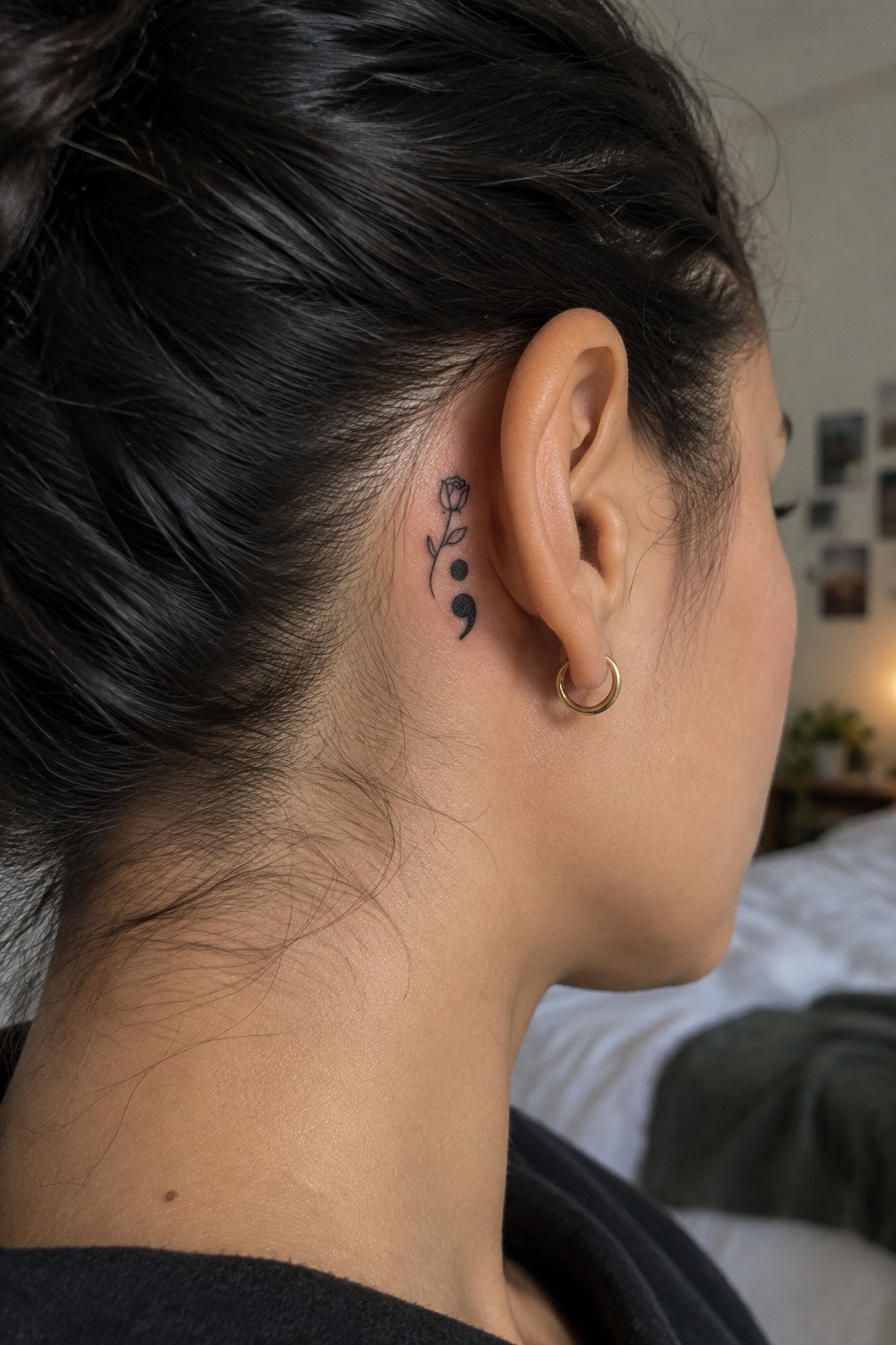 Small fine-line rose and semicolon tattoo behind a woman’s ear, above a gold hoop earring.