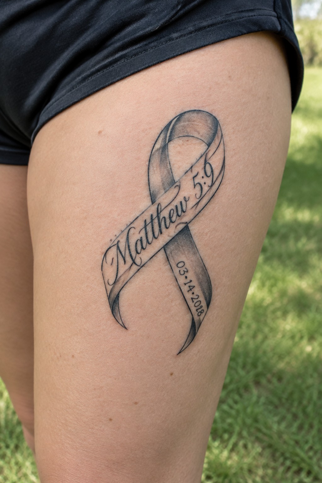 Shaded ribbon tattoo on the thigh with Matthew 5:9 and a date underneath