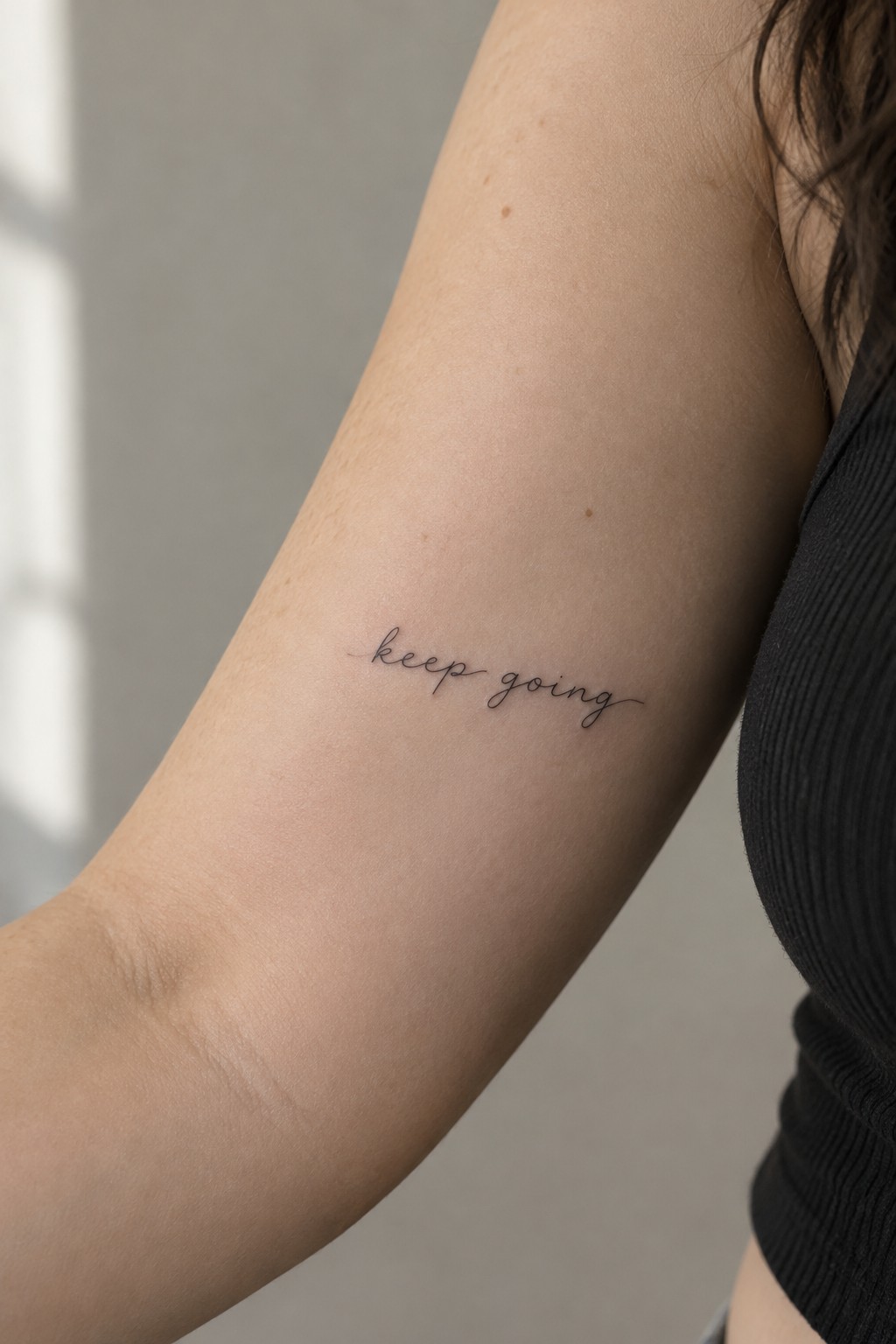 Small cursive “keep going” tattoo on the inner upper arm.