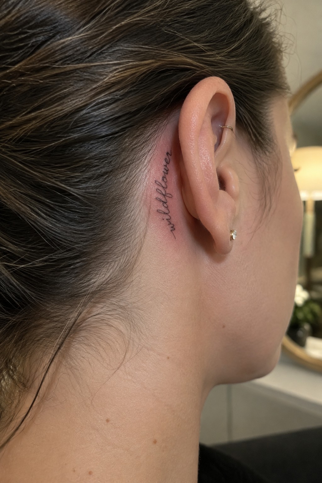 Fine-line “wildflower” script tattoo behind the ear, next to a small ear piercing.
