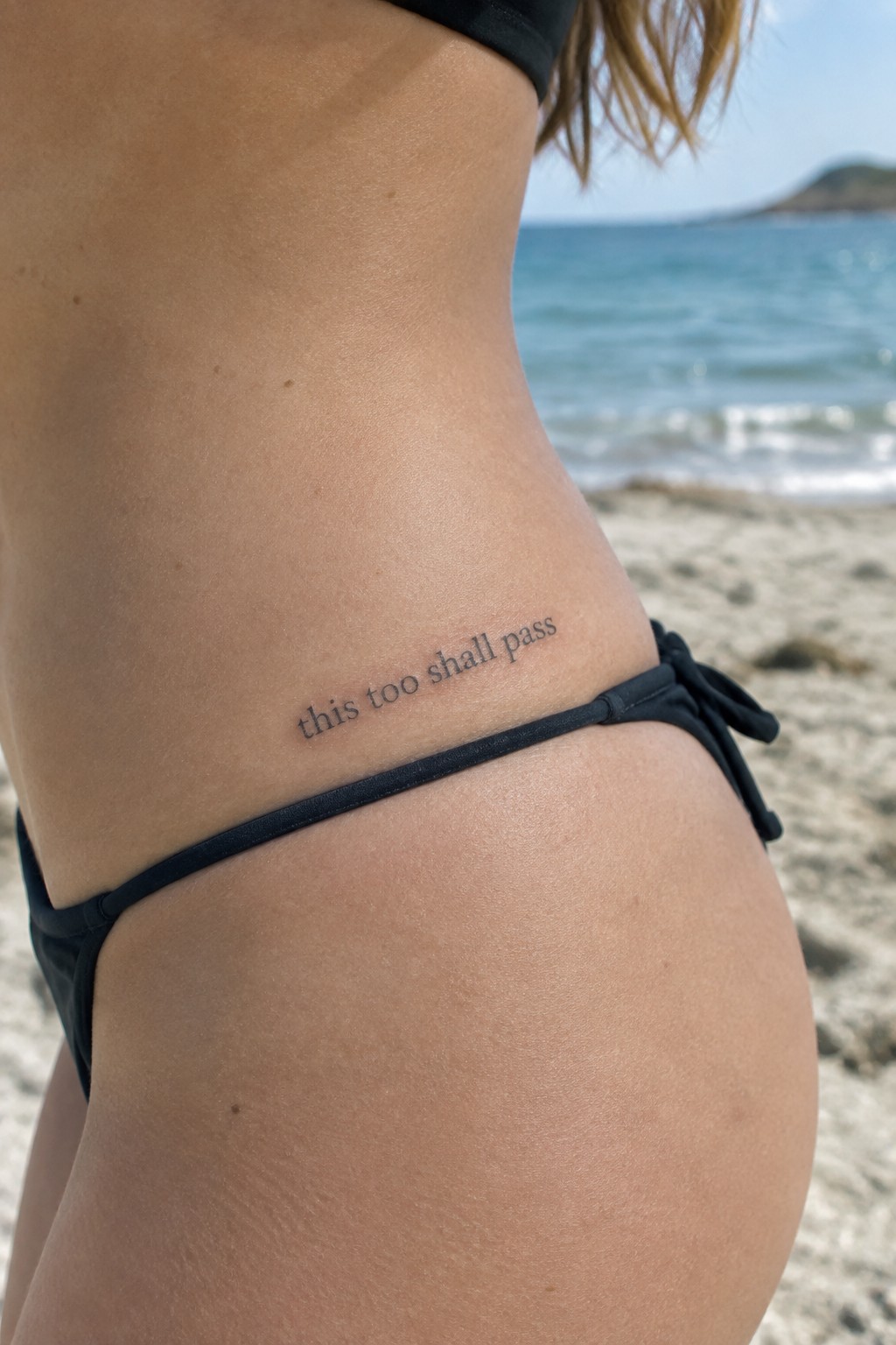 Small serif script tattoo reading “this too shall pass” on the side of a woman’s hip above a black bikini bottom.